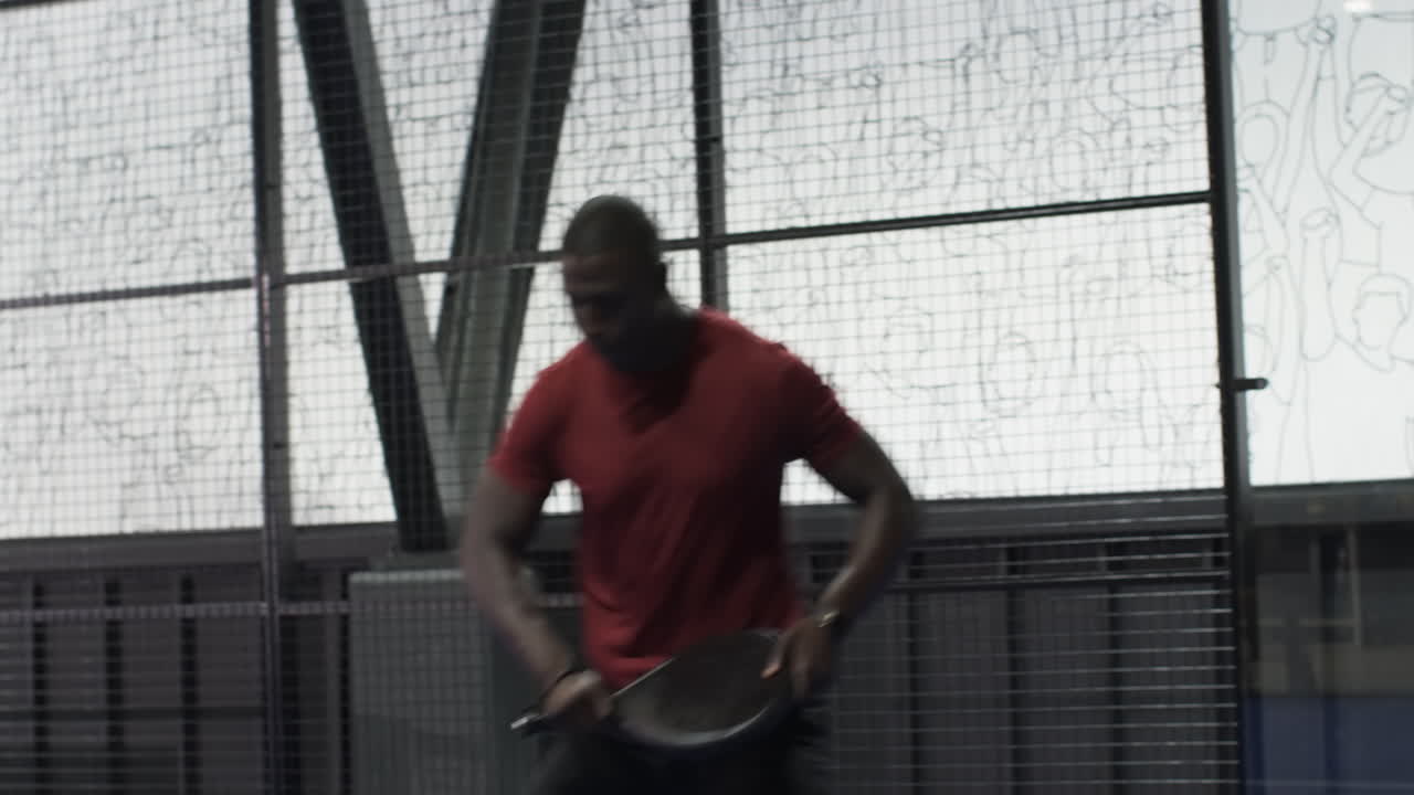 Mand spiller padeltennis, fokuserer på at slå bolden mod glasvæggen på en indendørs bane.
