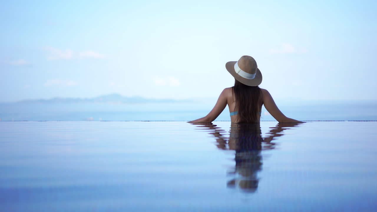 Eine Frau mit dem Rücken zur Kamera blickt vom Rand eines Infinity-Pools eines Resorts auf eine bergige Insel in tropischen Ozeangewässern.
