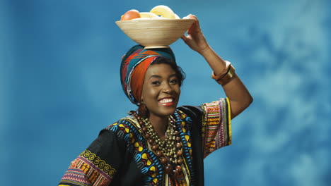 joven afroamericana alegre con turbante y ropa tradicional sonriendo alegremente a la cámara mientras sostiene un plato con frutas en la cabeza