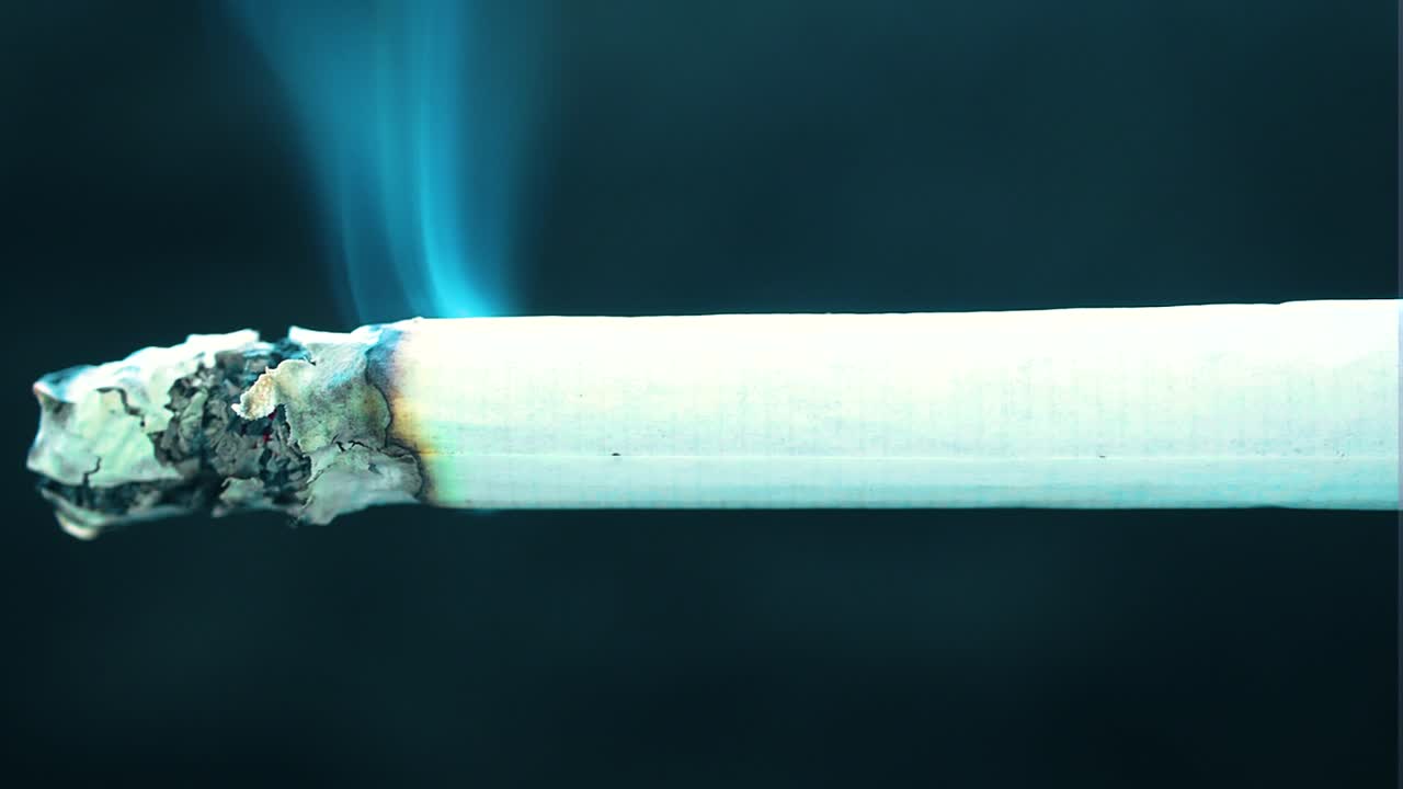 almost full isolated burning tailor made cigarette placed across the whole shot in the middle from one end to another ash is slowly falling down consuming paper releasing blue smoke black background