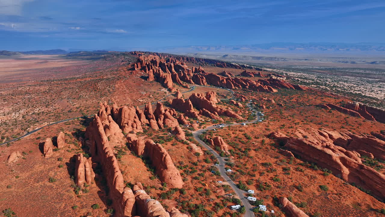 Footage over the multiple rocks with rounded tops and slopes. A highway with numerous campervans by its side along the mountains. Aerial perspective on the Arches National Park, Utah, USA