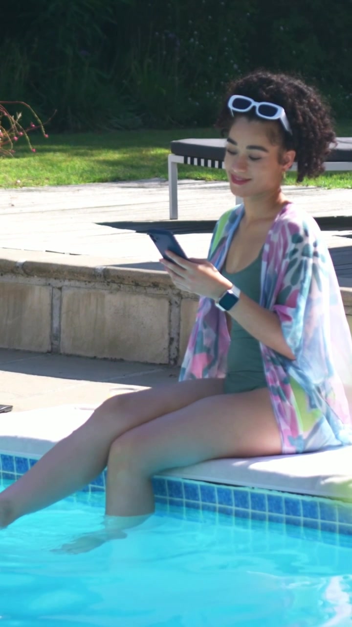 Vertical video: Holding tablet, woman in teal swimsuit relaxing at pool edge scrolling, sunglasses