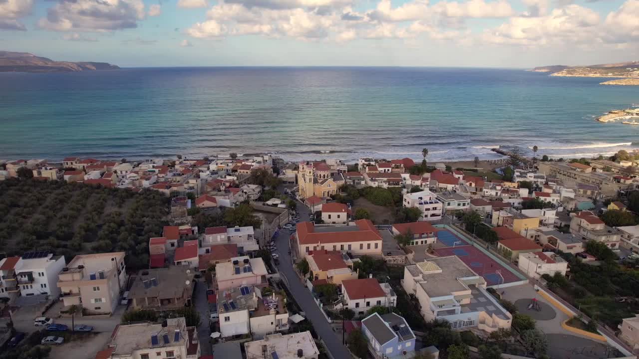 Aerial view of coastal village with red-roof houses near calm sea and olive groves. Rolling waves, cloudy sky, and distant mountains complete scenic and peaceful Santorini landscape.