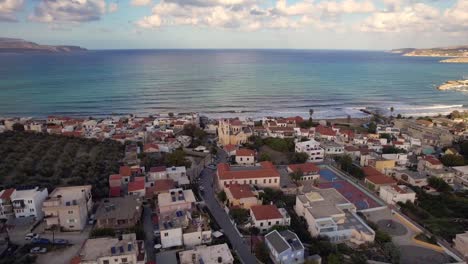 aerial view of coastal village with red-roof houses near calm sea and olive groves. rolling waves, cloudy sky, and distant mountains complete scenic and peaceful santorini landscape.