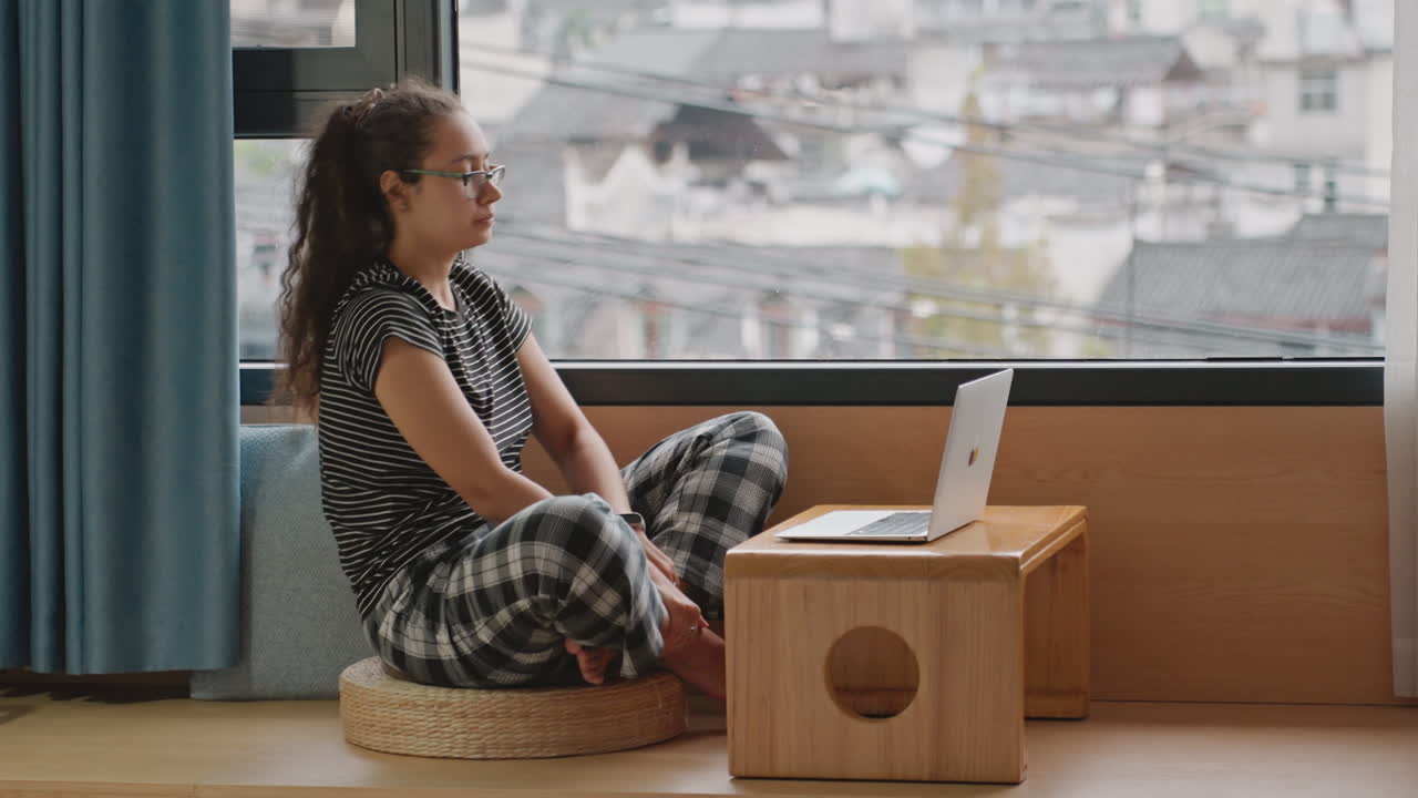 Hotel window student typing on laptop, young learner in pajamas seated on cushion at low table, city rooftops visible through glass, focused study session with headset nearby, natural light, calm