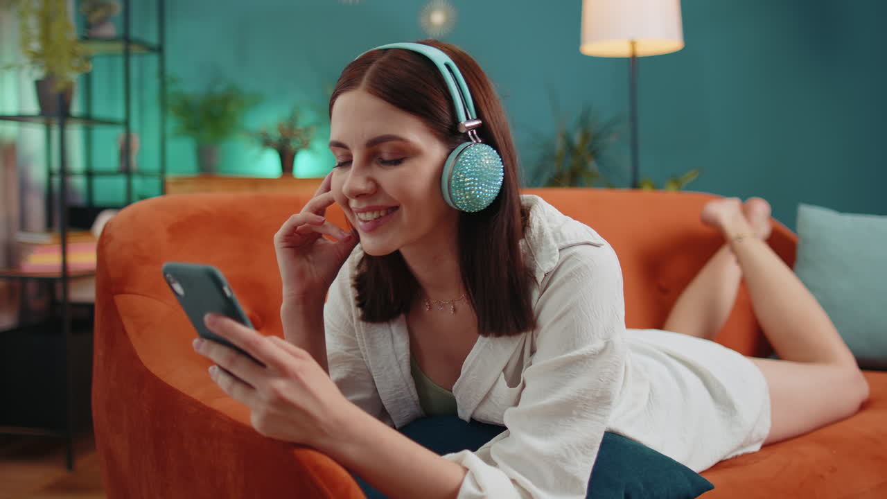 Giovane donna felice con le cuffie wireless che si rilassa sdraiata sul divano a casa ascoltando la sua musica preferita