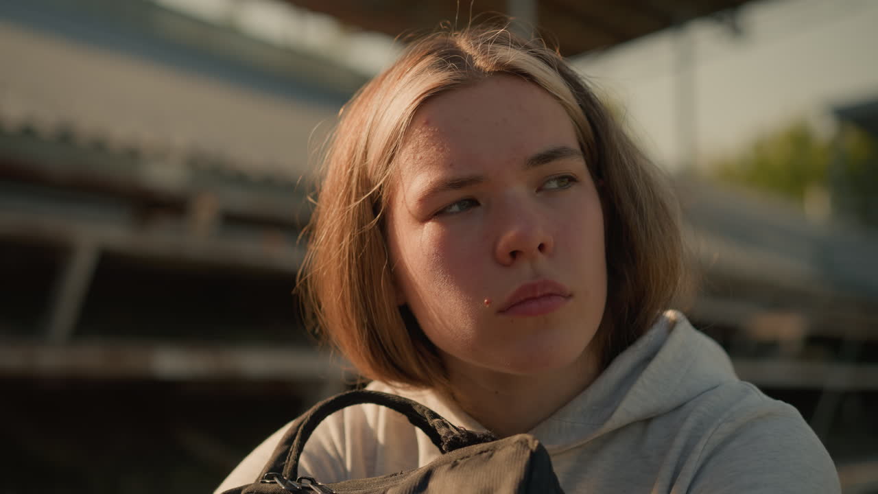 Close-up of lady in deep thought seated alone on empty stadium bleachers, hugging her bag with a reflective expression and distant gaze, sunlight softly illuminates her
