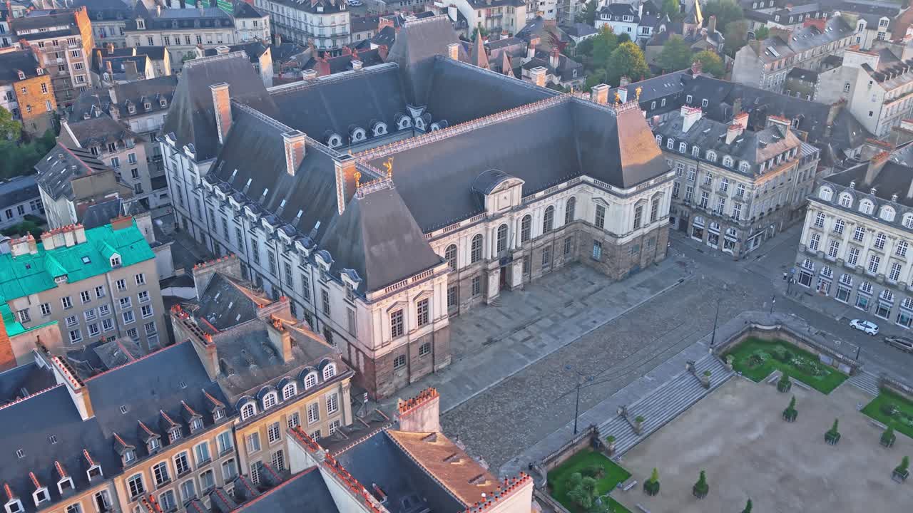 Diagonal drone approach toward the Parlement de Bretagne at sunrise, with a tilt-down revealing the square, rooftops, trees and surrounding streets in Rennes - Brittany
