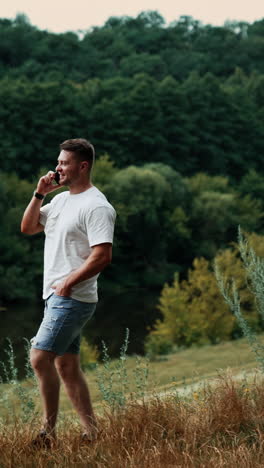 relaxed caucasian man walks by the grass speaking on the phone. man working remote having business conversation in nature. vertical video