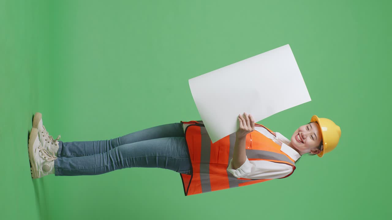 Full Body Side View Of Asian Female Engineer With Safety Helmet Looking At Blueprint In Her Hands While Standing In The Green Screen Background Studio