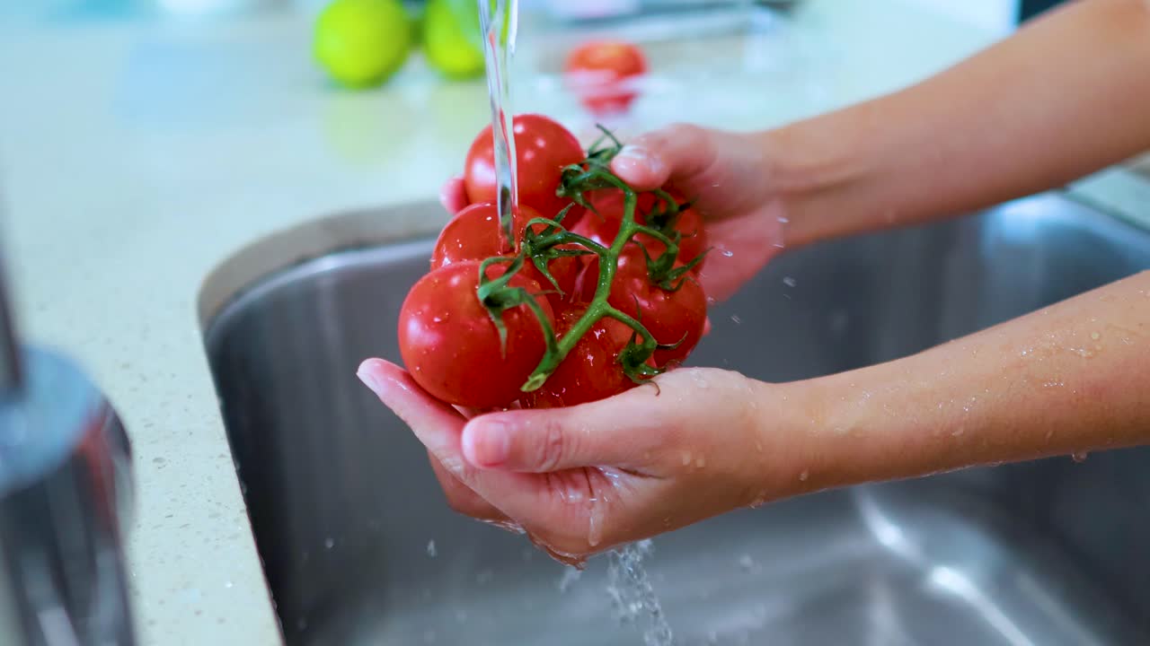 Hands rinse a cluster of ripe vine tomatoes under running water in a bright kitchen, with natural lighting and close-up camera movement over a stainless steel sink
