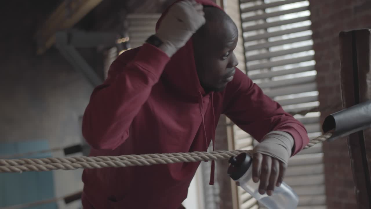 Black Boxer in Hoodie Leaning on Ropes and Drinking Water