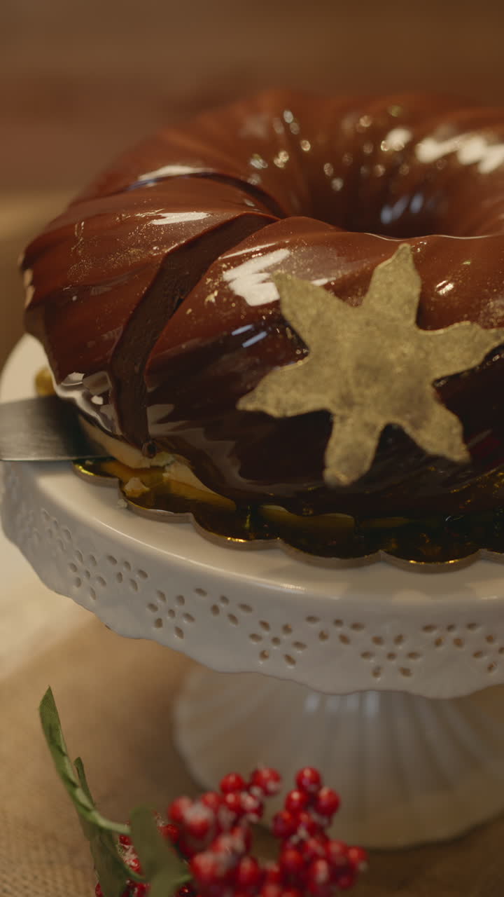Close-up of an elegant Christmas dessert with a glossy chocolate glaze, decorated with a gold star and presented on a white pedestal. The clip shows a cut slice