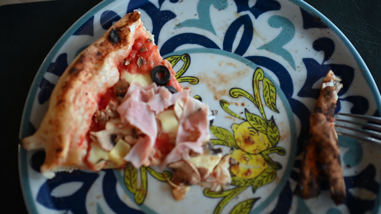 Fetta di pizza parzialmente mangiata con prosciutto, funghi, olive e formaggio su un piatto decorativo.