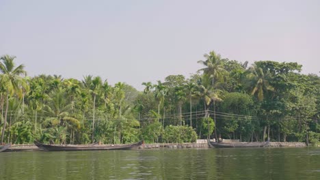 Moviéndose-Lentamente-A-Lo-Largo-De-Los-Remansos-De-Kerala-Mostrando-Barcos-De-Transporte-Tradicionales-Amarrados-En-La-Orilla-Con-Palmeras-Detrás