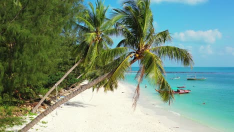 Tall-palm-trees-bent-over-white-sand-of-exotic-beach-washed-by-peaceful-turquoise-lagoon-with-anchored-fishing-boats-in-Thailand