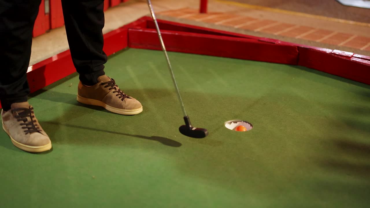 A player putts an orange ball on a mini-golf course. Indoor setting with artificial lighting and a focus on precision