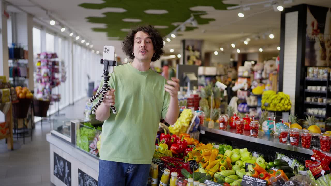 A brunette guy with curly hair blogs about food in a supermarket and films himself on camera using a smartphone