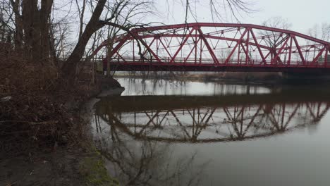 A-small,-picturesque-arched-red-steel-bridge,-as-seen-from-a-little-park,-crossing-a-small-river-in-upstate-New-York-on-a-rainy,-dreary,-early-spring-day