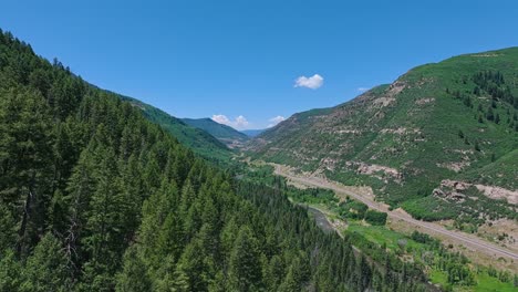 Flying-along-the-Slate-River-and-a-forested-valley-with-a-road-through-the-middle-near-the,-Colorado,-USA