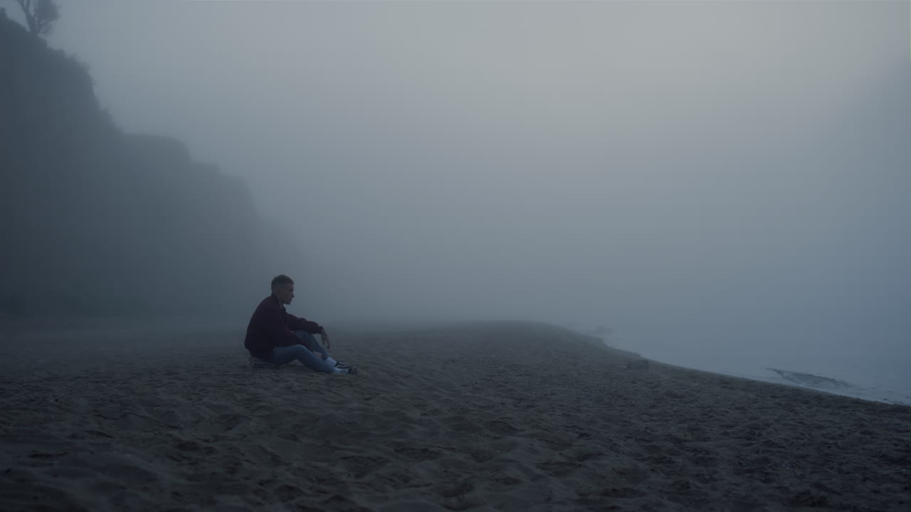 Depressed guy sitting sea beach in foggy morning. Worried man touching sand