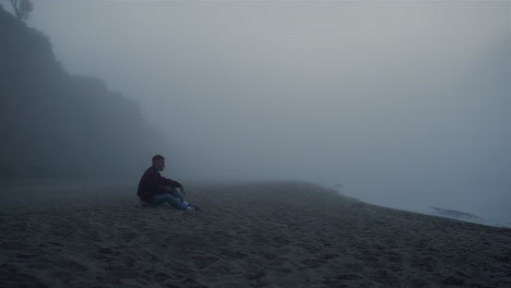 depressed guy sitting sea beach in foggy morning. worried man touching sand