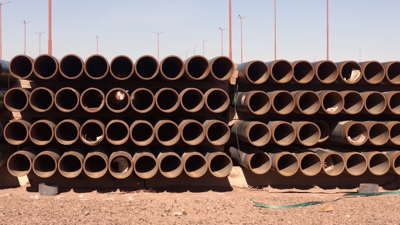 Sporende opptak fra bakkenivå som passerer rader med stablede stålrør brukt til olje- og gassboring og rørledningsinfrastruktur på et tørt industriområde