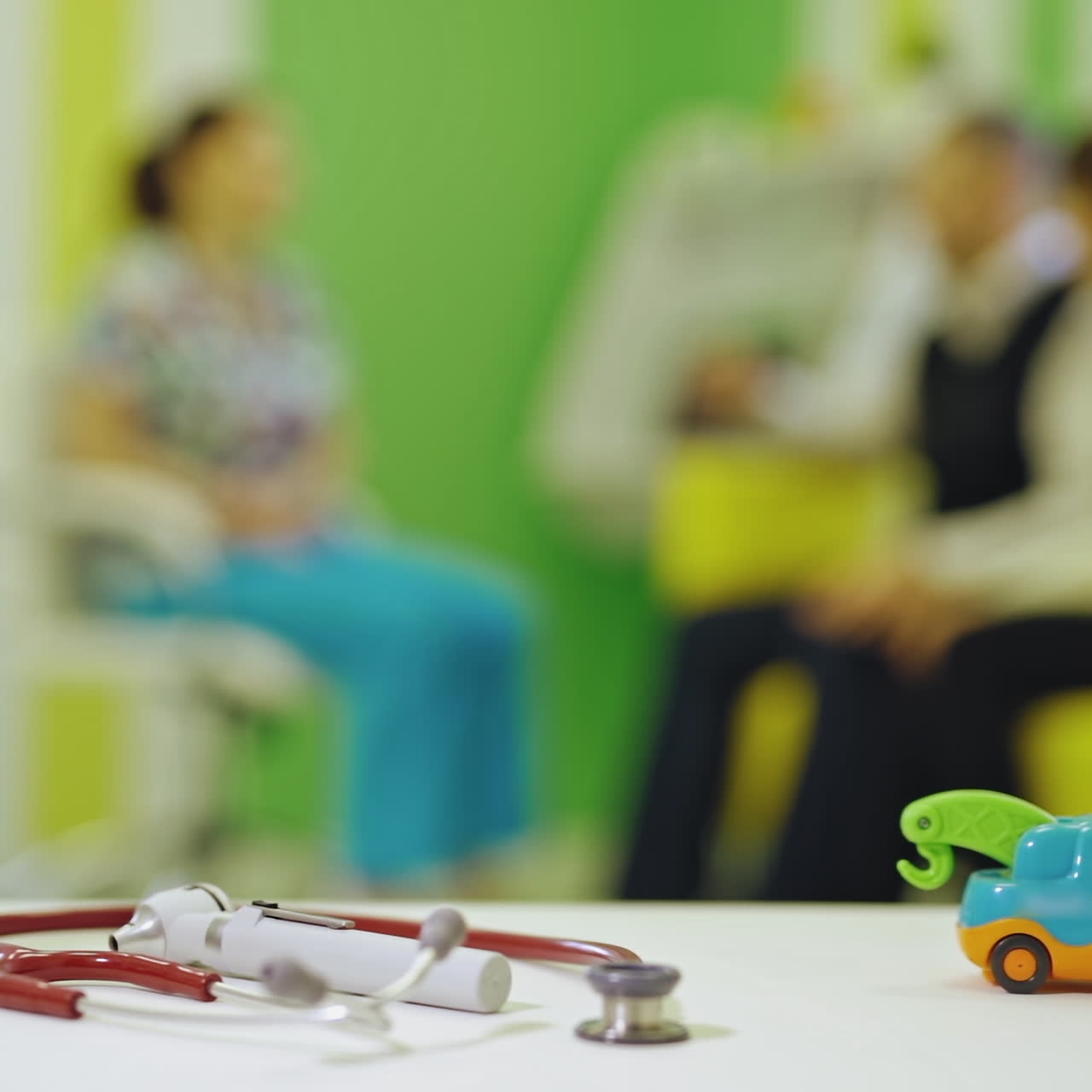 Thermometer, phonendoscope and a toy car are on the table indoors. Toy and medical equipment on the background of parents sitting against the doctor in the room.