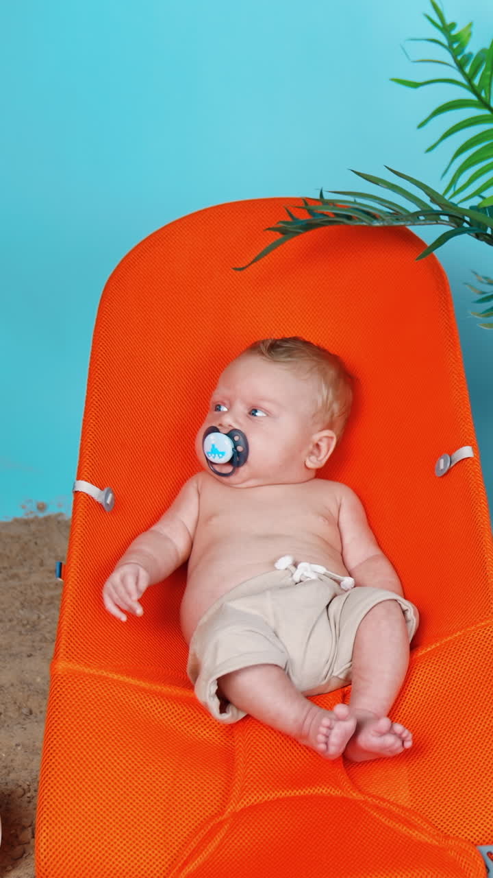 Sweet little baby in shorts and with pacifier lies on the orange chair. Beach sand and plant pot around the kid. Vertical video