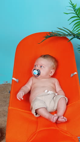 sweet little baby in shorts and with pacifier lies on the orange chair. beach sand and plant pot around the kid. vertical video