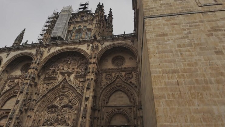 Ripresa a mano che si innalza sulla facciata gotica ornata e sull'imponente campanile dell'iconica Cattedrale Nuova di Salamanca. Cielo drammatico e coperto, intricata lavorazione in pietra, angolazione dinamica