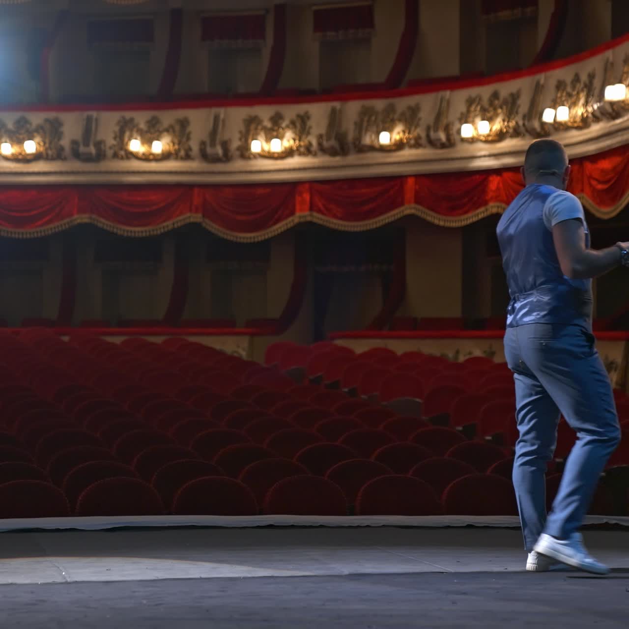 Man in front of empty theatre hall with red velvet chairs. Actor is standing on stage and talking with gestures before the performance