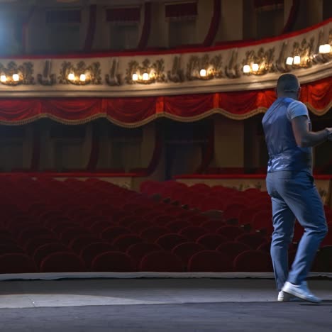 man in front of empty theatre hall with red velvet chairs. actor is standing on stage and talking with gestures before the performance