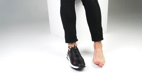 male sitting on a white cube with one foot in a shoe and another is bare. man moving feet on a white backdrop.