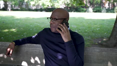 un hombre negro calvo adulto con gafas está escuchando en un teléfono inteligente mientras está sentado afuera en un parque en un día soleado