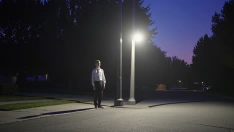 young man in a white shirt and dark pants stands beneath a brightly lit streetlight in a quiet residential area, the dusky sky hinting at the approaching night