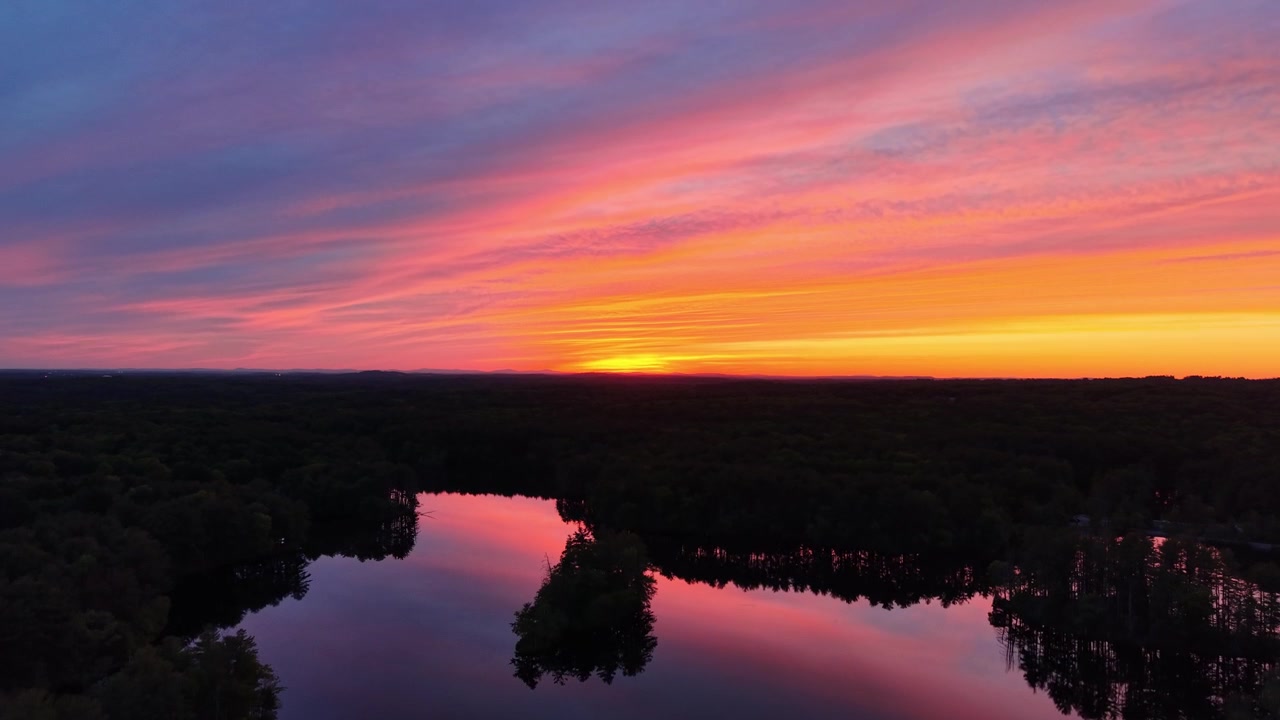 Droneudsigt over farverig pink og orange solnedgangshimmel, der spejler sig i mørkt søvand omgivet af skov i Harold Parker State Forest, Massachusetts.