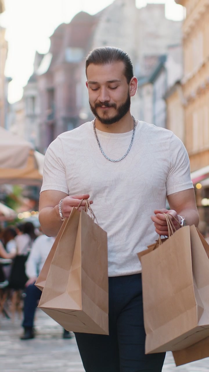 Felice giovane uomo, acquirente compulsivo, dopo i saldi, con borse piene, che cammina per la strada in città.