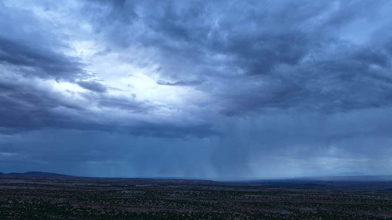 4K drone-timelapse som fanger en stemningsfull blåtime-skumring over en vidstrakt mesa i sørvest. Regnbyger faller fra mørke, tunge stormskyer som har et mykt, eterisk skjær midt på himmelen.