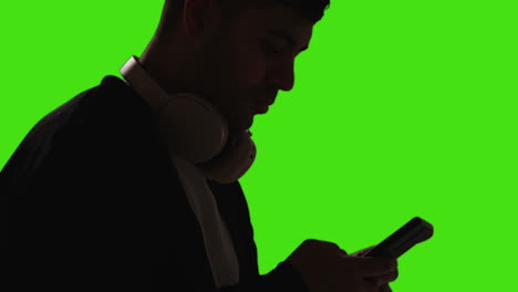 Close-Up-Of-Man-Wearing-Wireless-Headphones-Text-Messaging-On-Mobile-Phone-Against-Green-Screen-With-Low-Key-Lighting-1