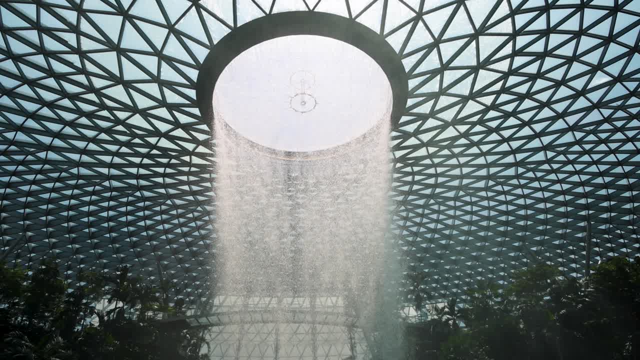 A dramatic indoor waterfall pours from a circular skylight in a geometric glass dome, surrounded by lush greenery and illuminated by natural daylight