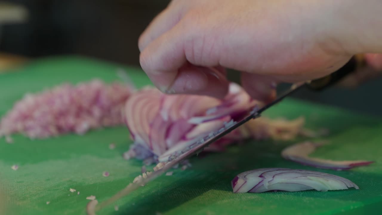 Close up, A chef slices a red Spanish onion with a very sharp black handled chef knife sliding it off the knife onto the green chopping board ready to finely dice