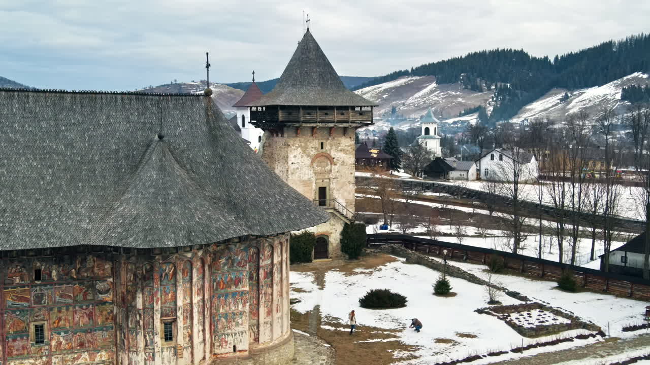 Aerial drone view of The Humor Monastery in winter, Romania. Main painted church and the Tower, yard with snow and tourists, bare trees, residential buildings
