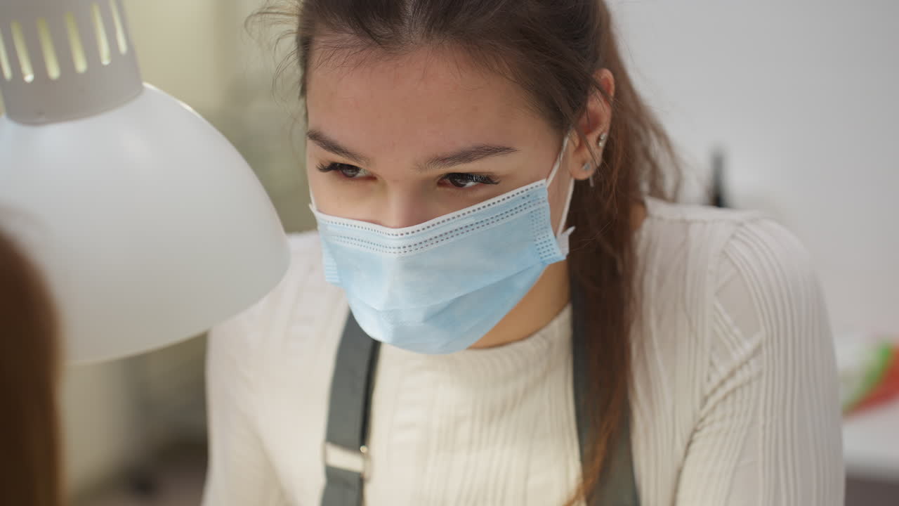 Closeup view of nail technician in face mask and gloves engaging attentively with client while performing manicure, highlighting focused expression and professional demeanor under salon lighting setup