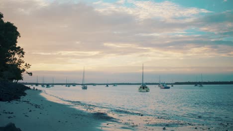 Dozens-of-small-boats-float-upon-the-surface-of-the-ocean-just-shy-off-the-shore-of-a-beautiful-beach-reflecting-the-suns-gorgeous-warm-glow-off-of-its-water-as-white,-puffy-clouds-sit-overhead