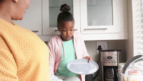 Happy-african-american-mother-and-daughter-washing-up-and-drying-dishes-in-kitchen,-slow-motion