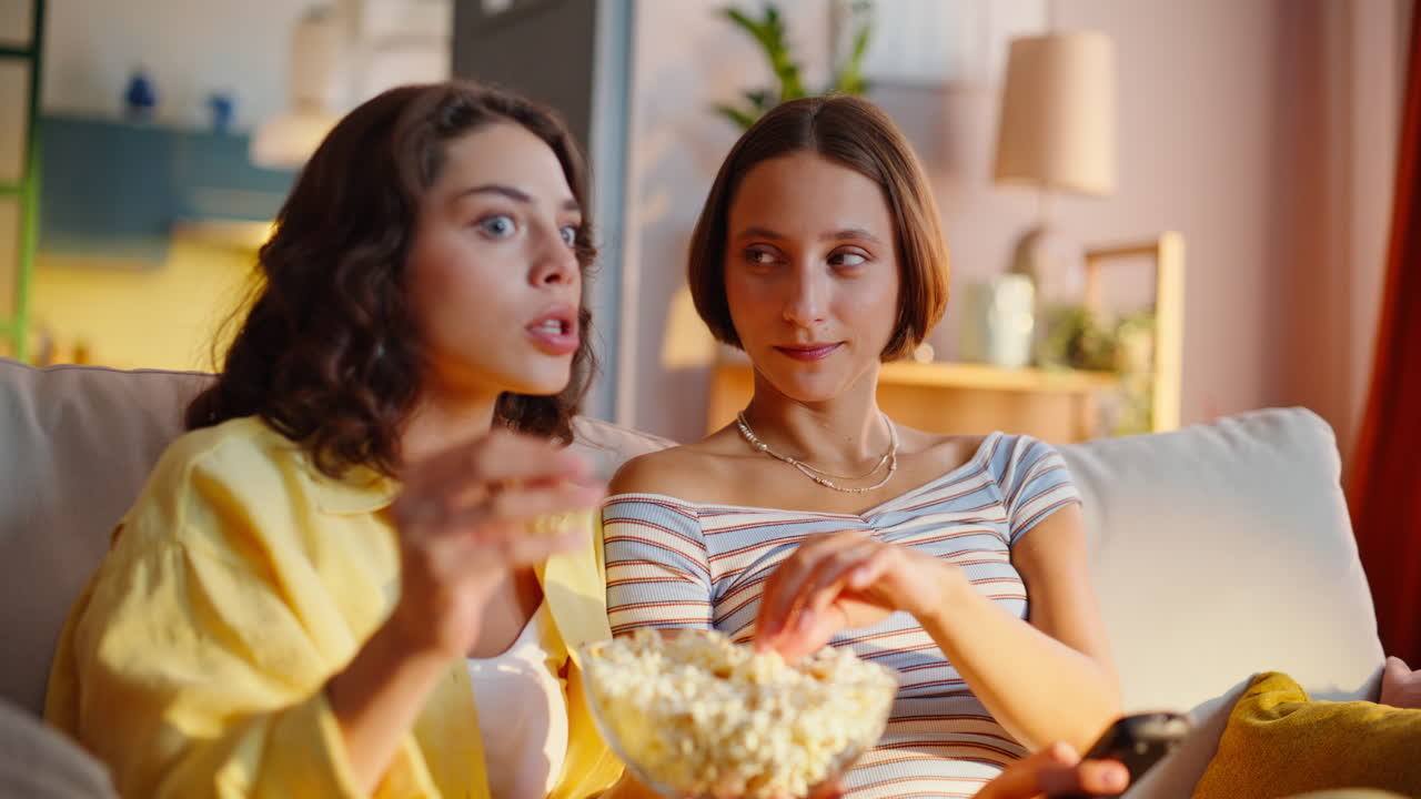 Afslappede søstre, der diskuterer film, mens de sidder hyggeligt i sofaen derhjemme med snacks i nærbillede