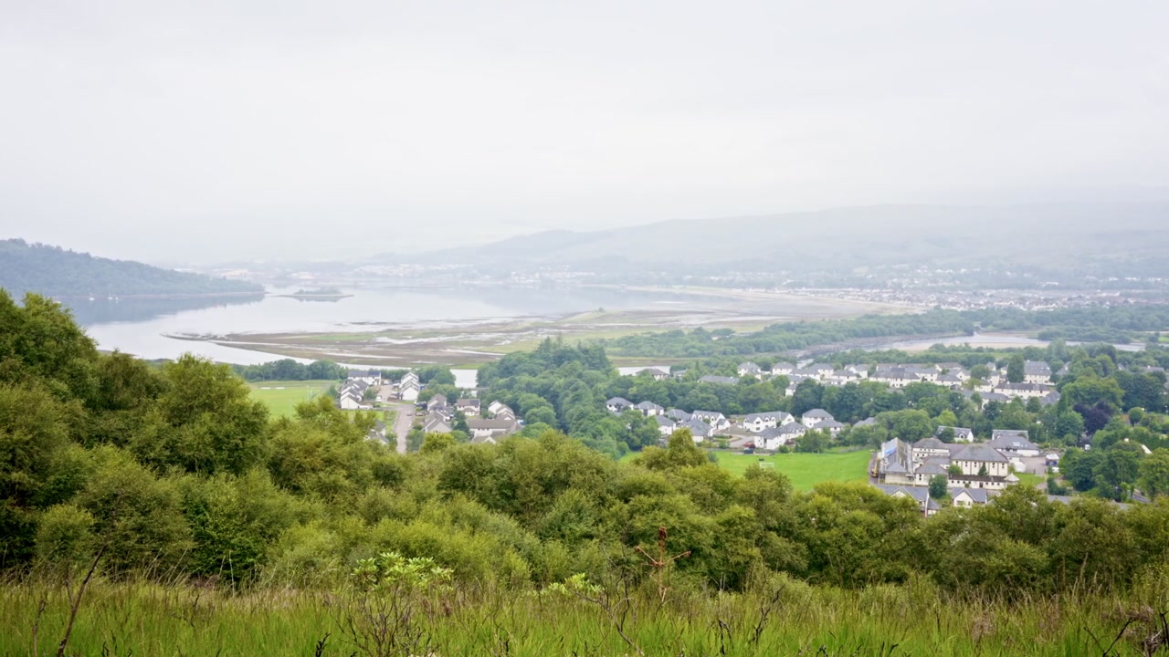 Scenic residential town nestled below lush green hills overlooking a broad misty sea loch. Trees and rolling terrain create a peaceful Highland landscape in Scotland.