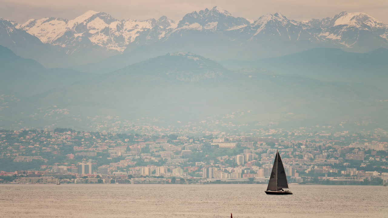 Antibes, France - May 6, 2025: Distant view of a sailboat moving on the sea with the city and the mountains on the background