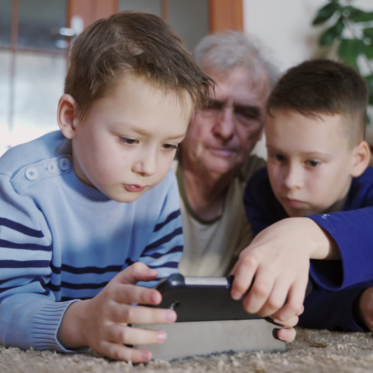 Two boys are playing on the tablets under the supervision of a grandfather in the room on the background of sofa and green plant on the weekday. Relatives.